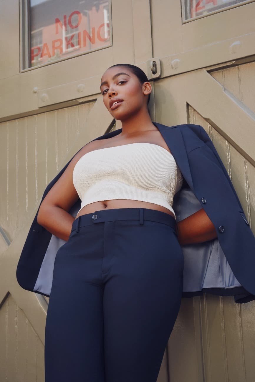 A woman casually leans against a wall, looking at the camera. Her navy blazer is draped over her shoulders.