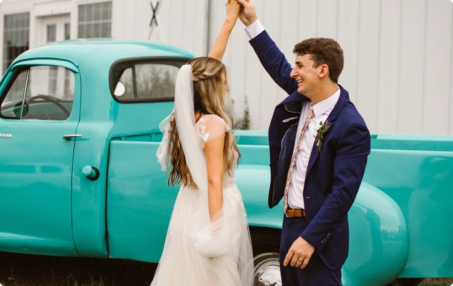 Groom in navy blue suit twirling bride in dancing wedding photo in front of teal truck.