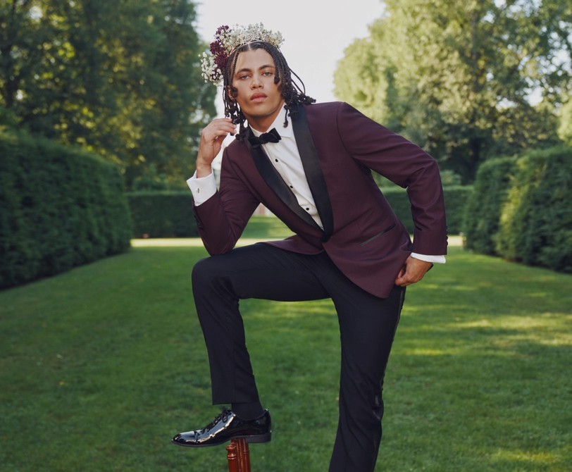 Prom king in a burgundy shawl lapel tuxedo and flower crown strikes a dramatic pose in the garden.