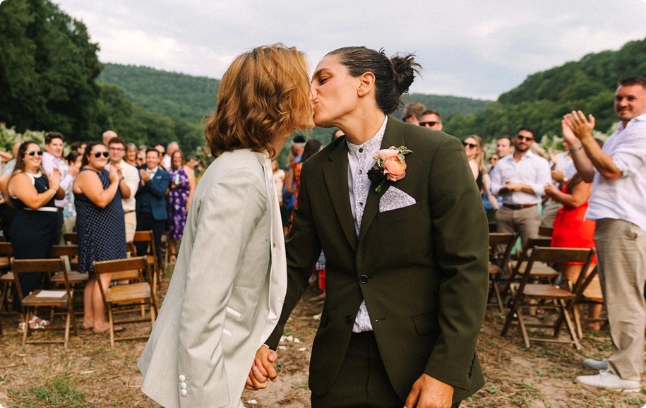 Two brides kissing in tan tuxedo and olive green suit & women's floral button-up at lesbian wedding.