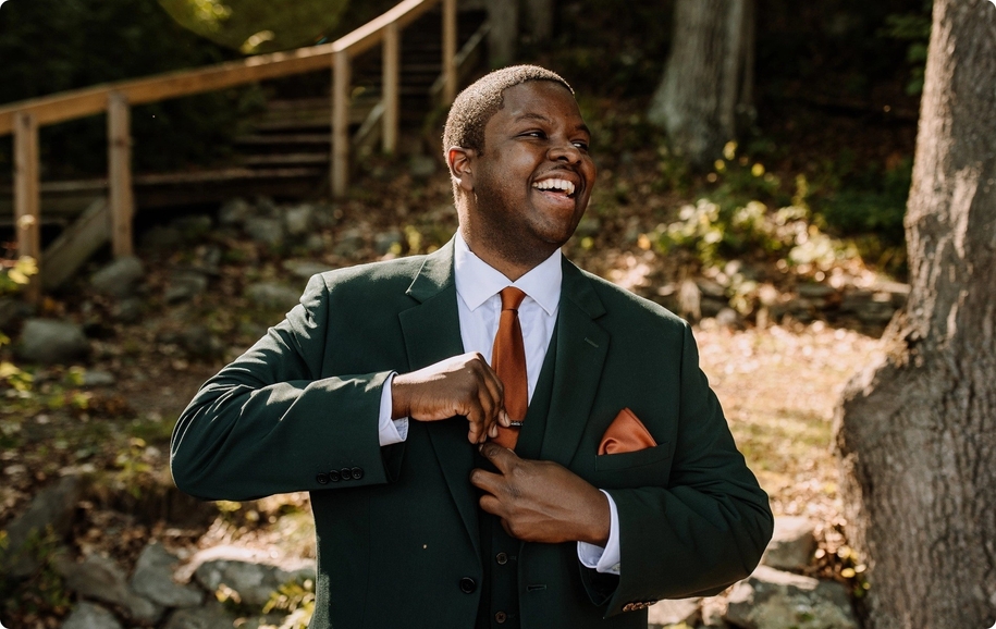 Dapper groom smiling in dark green 3 piece suit, copper pocket square, & burnt orange tie. 