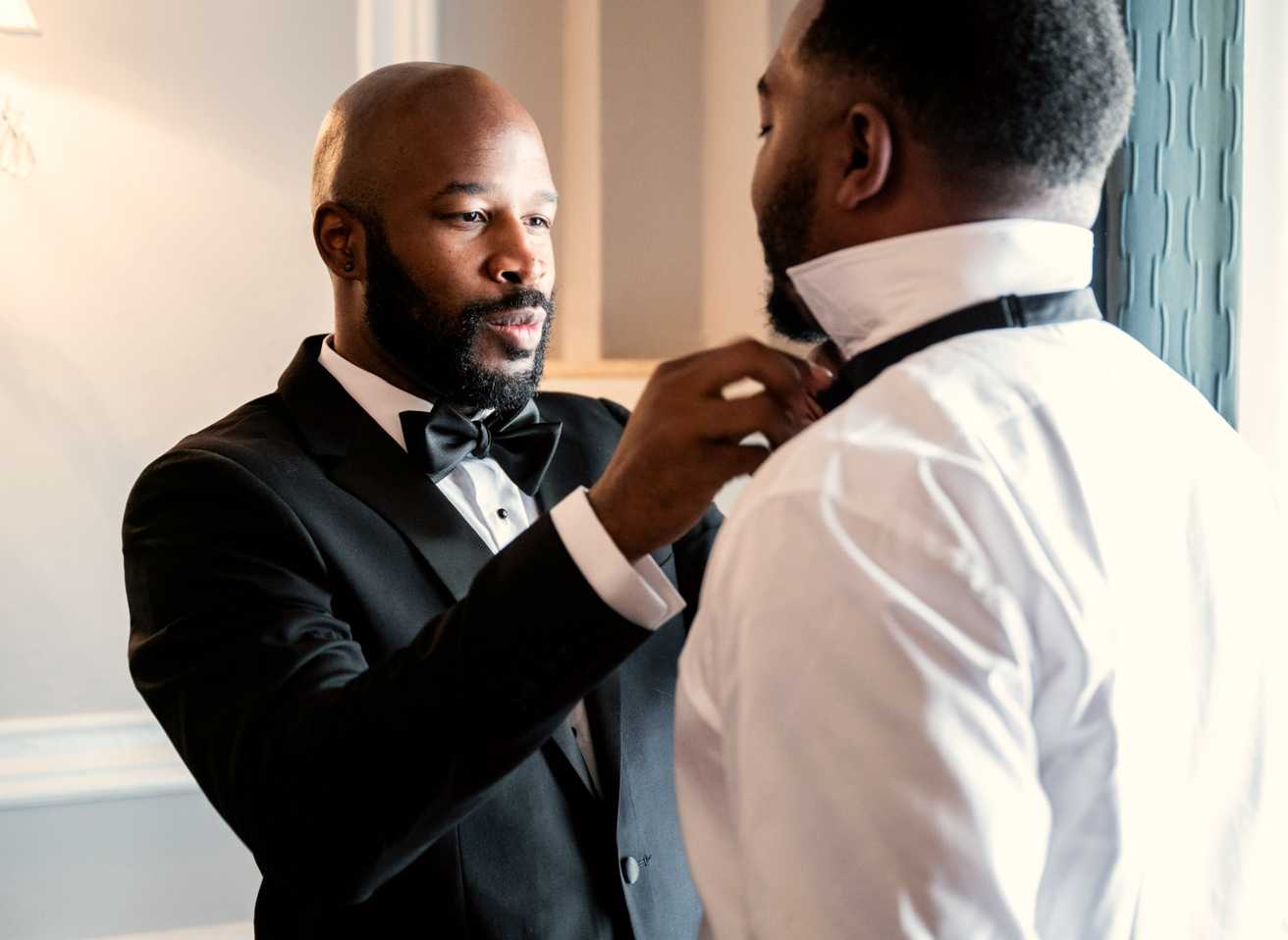 A man in a SuitShop tuxedo ties another man's bowtie while getting ready for a wedding.