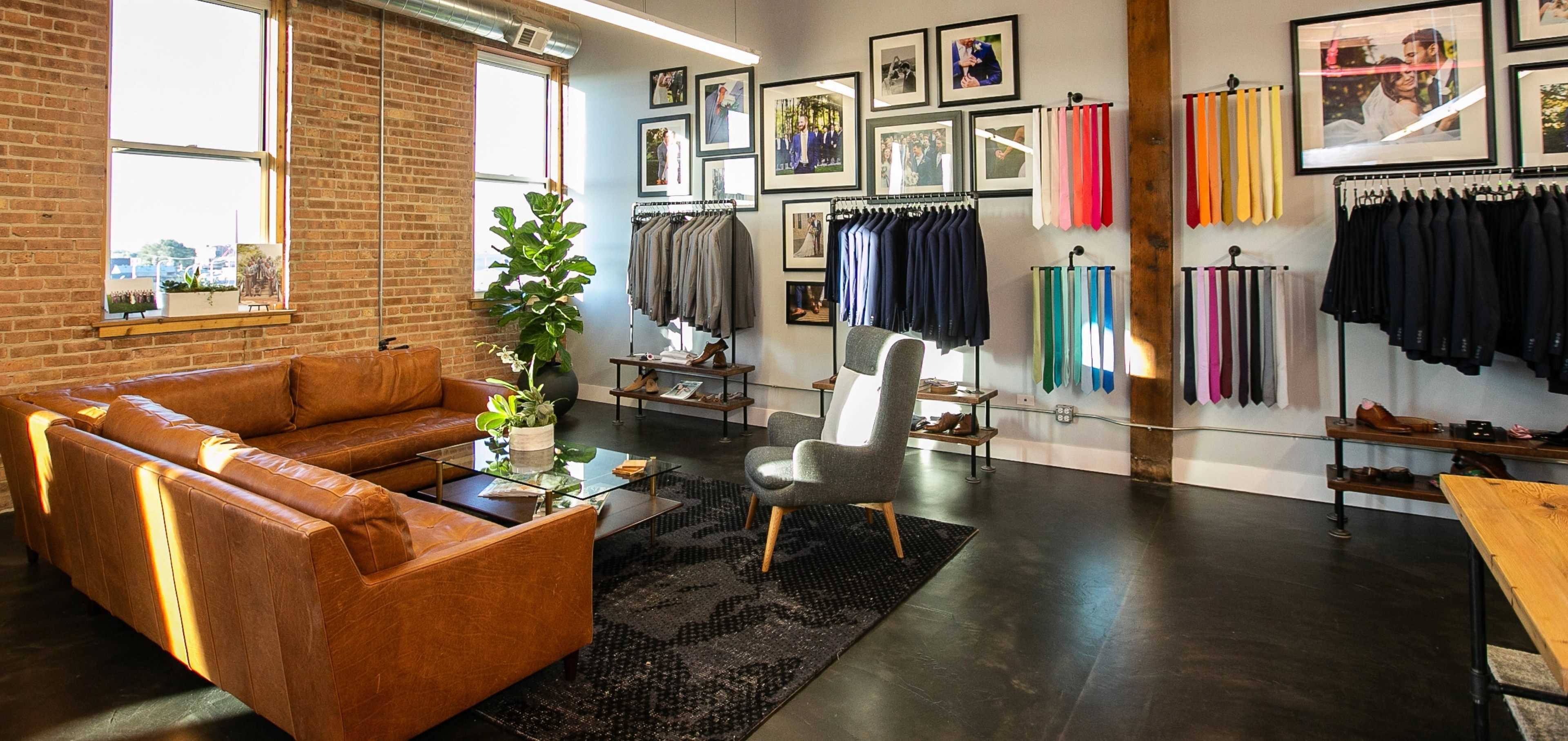 Interior of suit store with suit jackets, pants, shoes, colorful accessories, and lounge area.