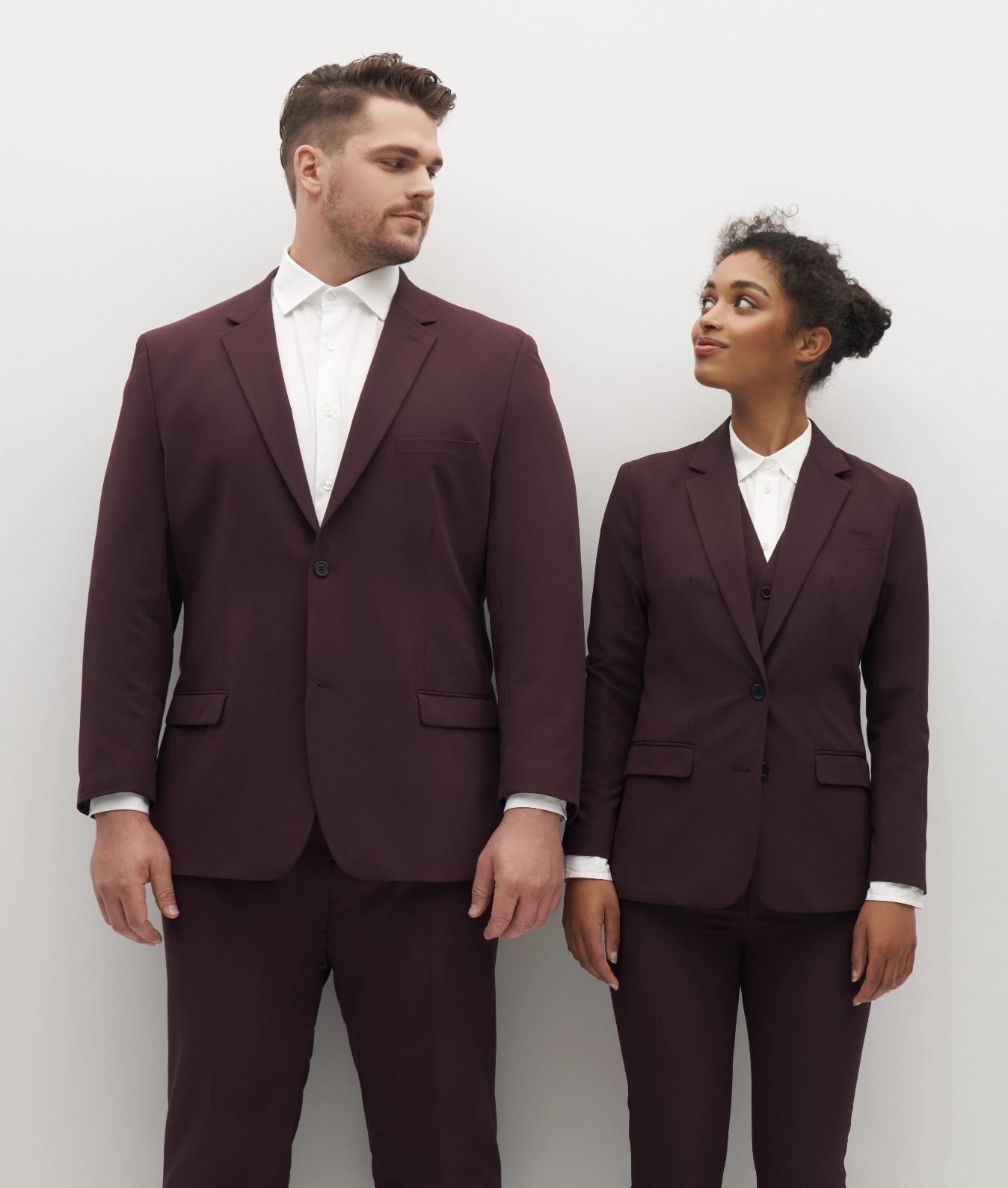 A large man and petite woman wear matching SuitShop burgundy jackets.